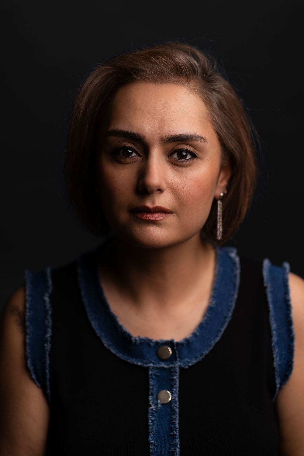 Portrait of a woman with short brown hair wearing a black and denim sleeveless top against a dark background.
