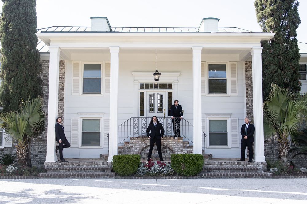 Four people posing in front of a grand white colonial mansion with tall columns and cypress trees.