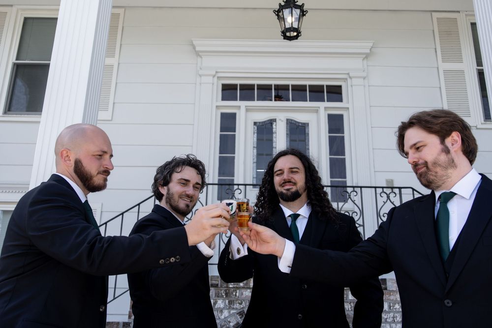 Four men in dark suits toasting with drinks outside a white house at a wedding celebration.