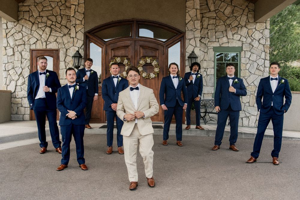 Groomsmen in navy suits and brown shoes show off their socks in a playful group photo at a stone-walled venue.