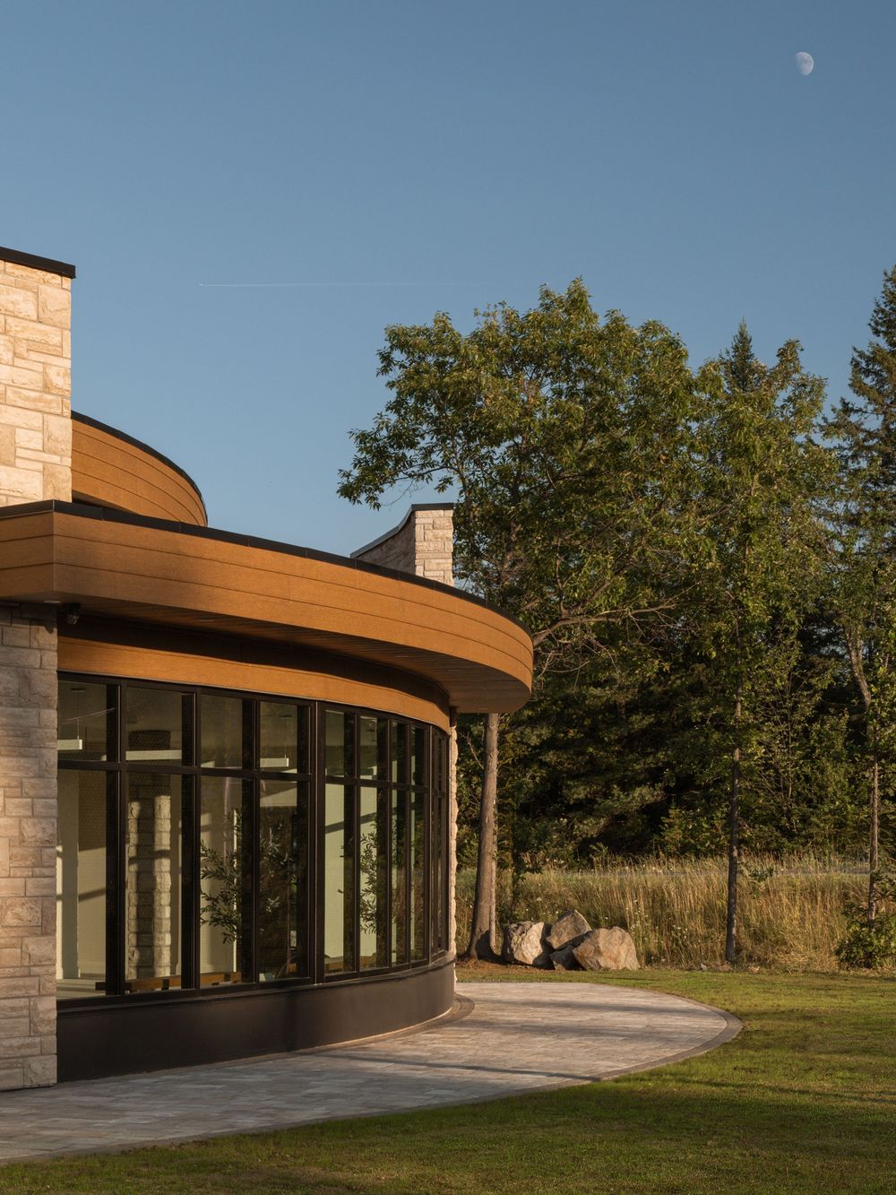 Curved modern building with wood soffit, stone facade, and floor-to-ceiling windows set against lush trees under a clear blue sky.