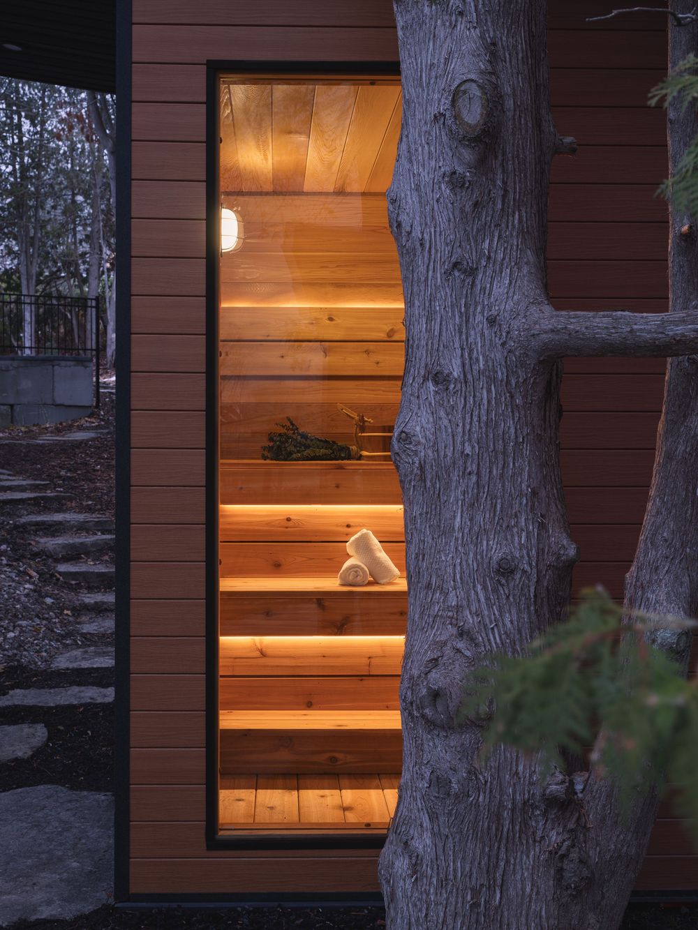 Warm glowing light shines through wooden slats of a rustic cabin window at dusk, with tree trunks silhouetted in foreground.