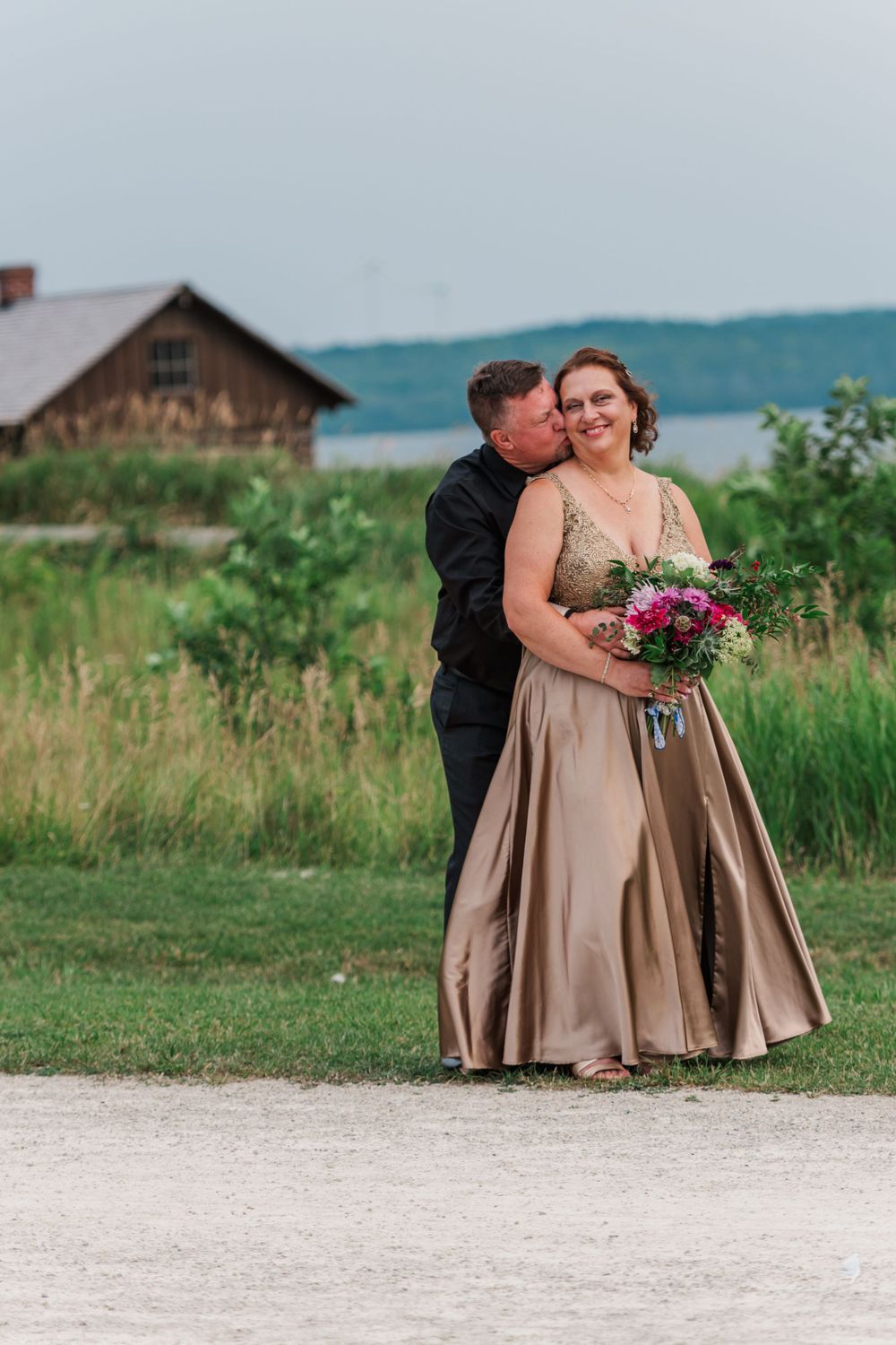 Couple in formal attire embrace in rural setting with rustic barn and hills in background. fayette state park