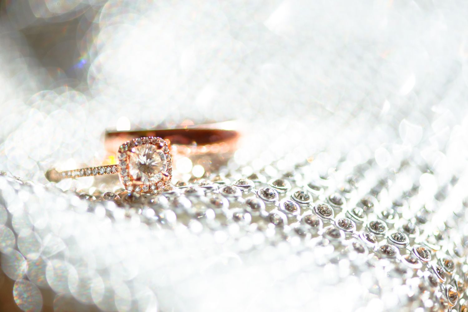 Rose gold engagement ring with diamond center stone rests on white lace fabric with sparkly rhinestone details.