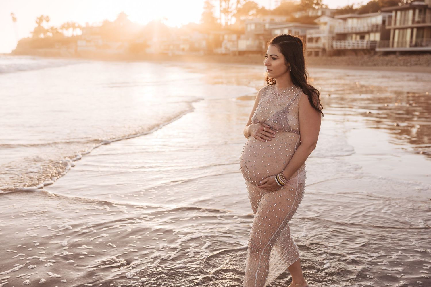 Expectant mother in a knit dress stands on beach at sunset while waves lap at the shoreline.