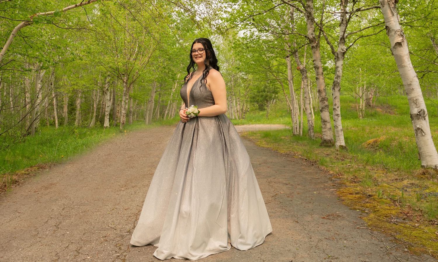 Young woman in elegant silver ombre ball gown posing on forest path surrounded by green birch trees.