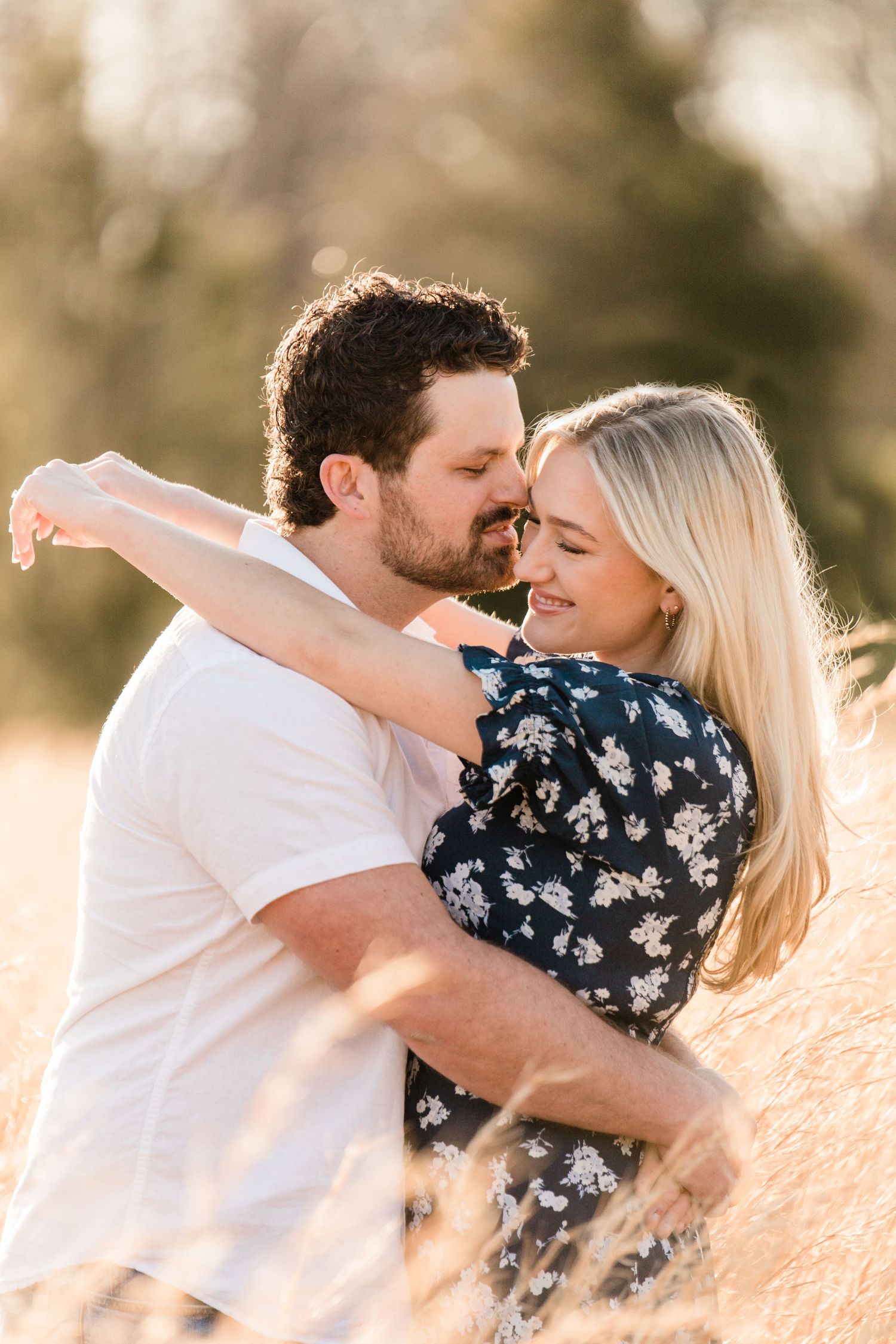 A romantic couple in white and navy floral embraces outdoors in warm golden sunlight.