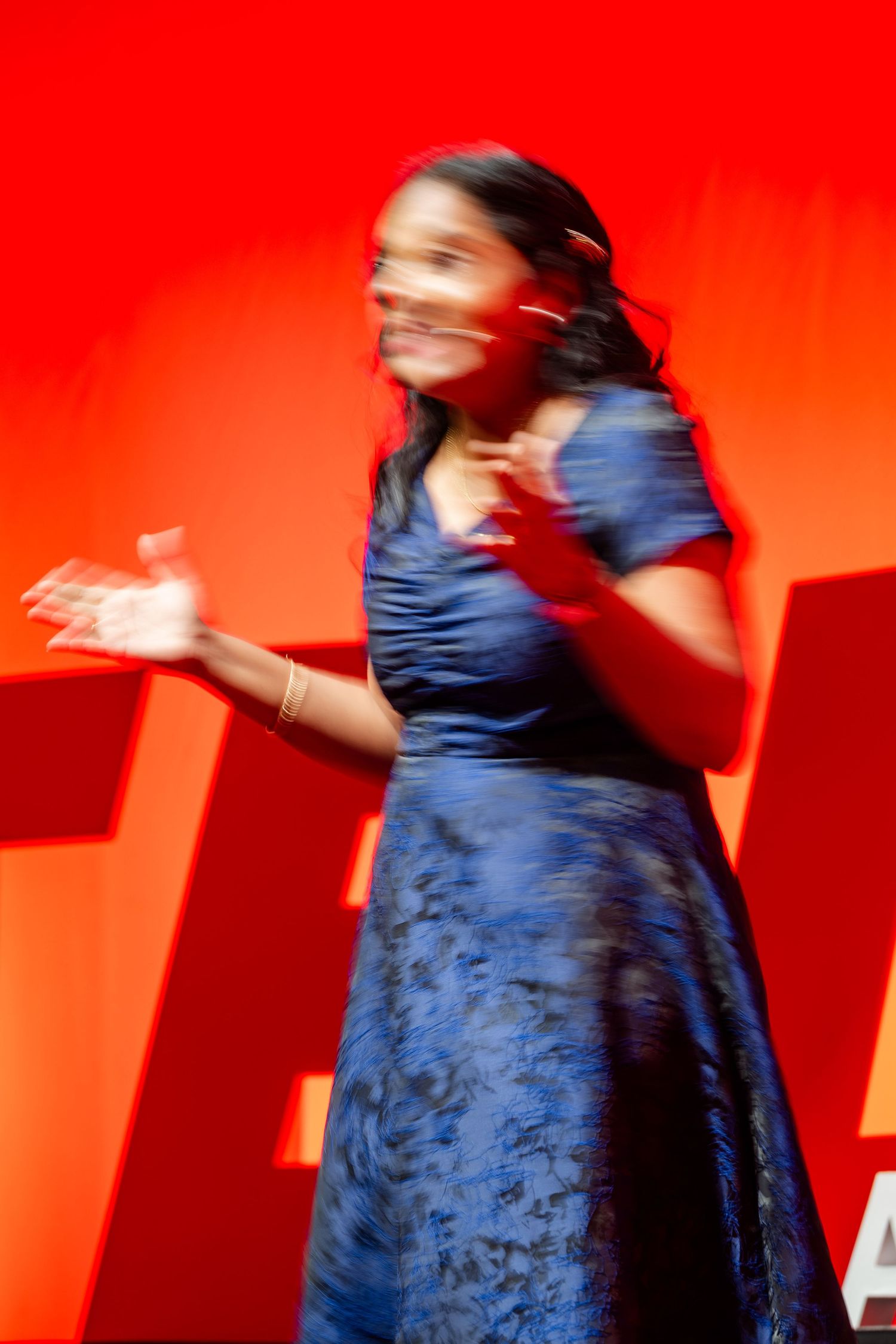 Motion-blurred figure in flowing blue dress gestures dramatically against vivid red background during stage performance.