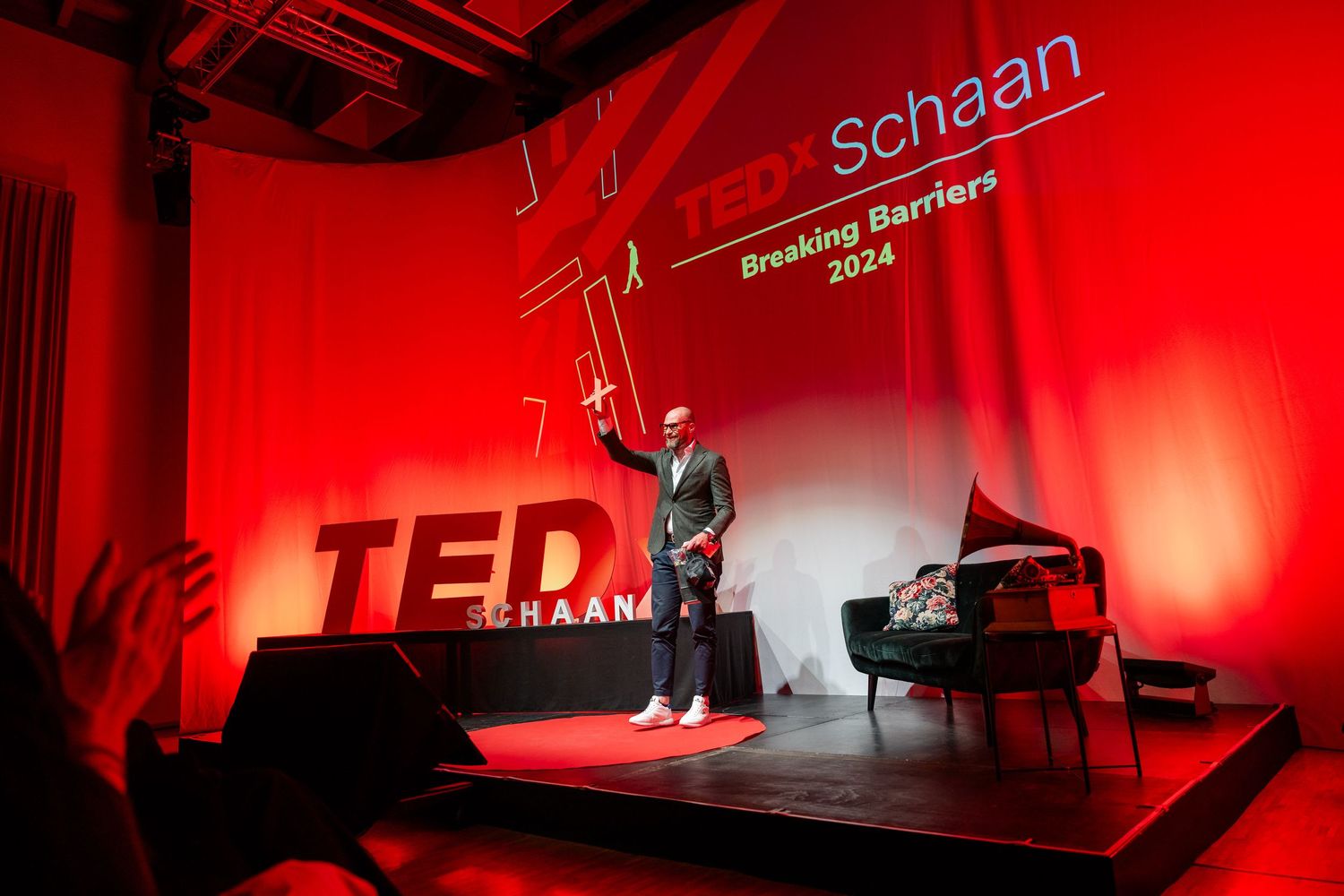 A speaker stands on a brightly lit red stage at a TEDx event with 'schaan' text visible above.