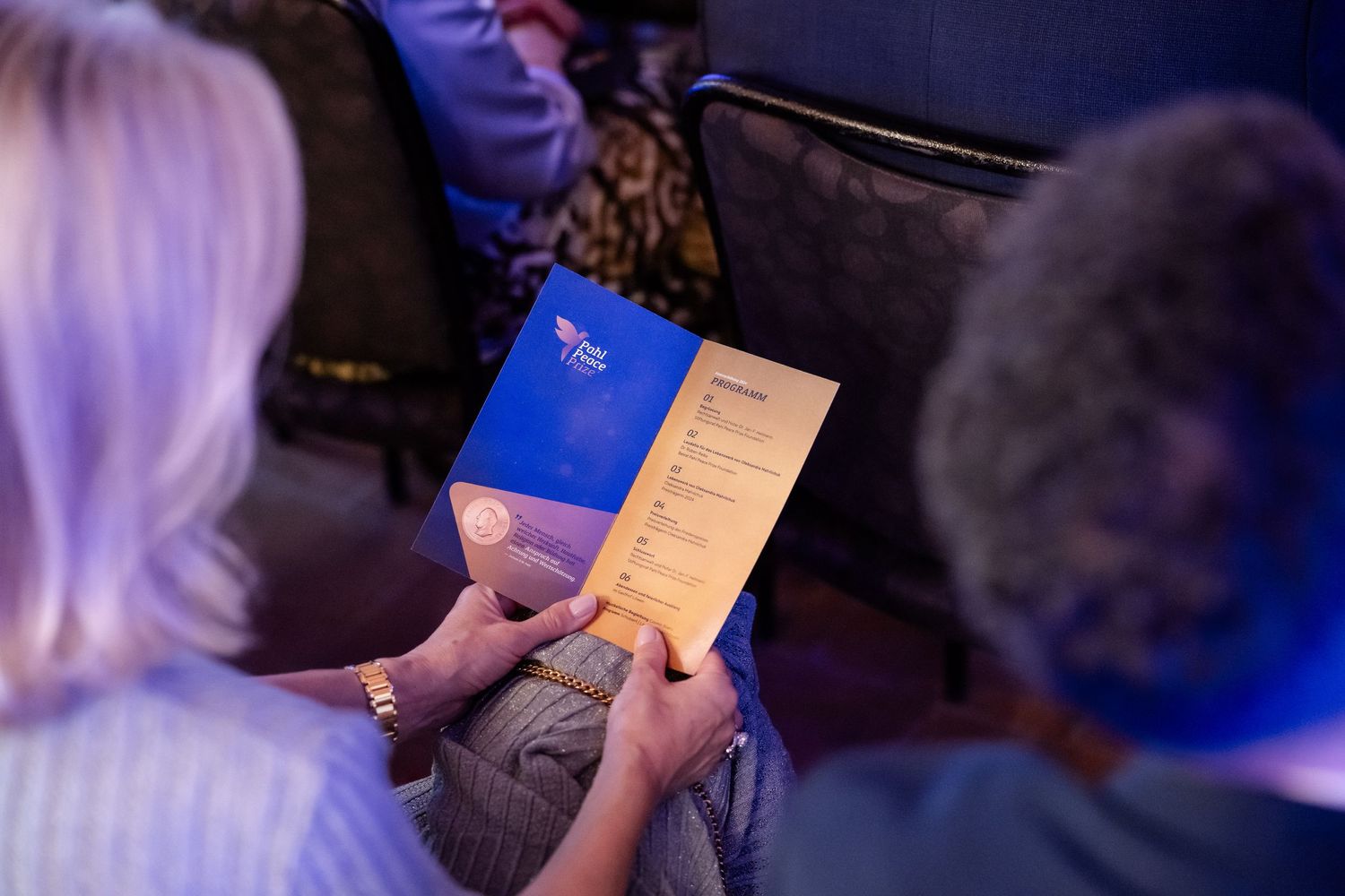 Event attendees reading a blue program or menu during a dimly lit performance or gathering.