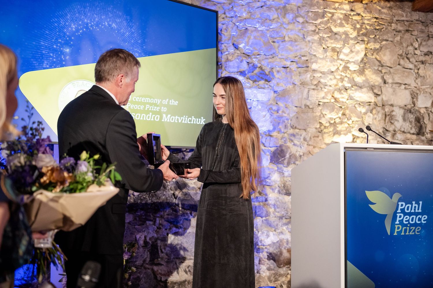 A person in a black dress receives an award on stage with a blue backdrop and stone wall during an event ceremony.