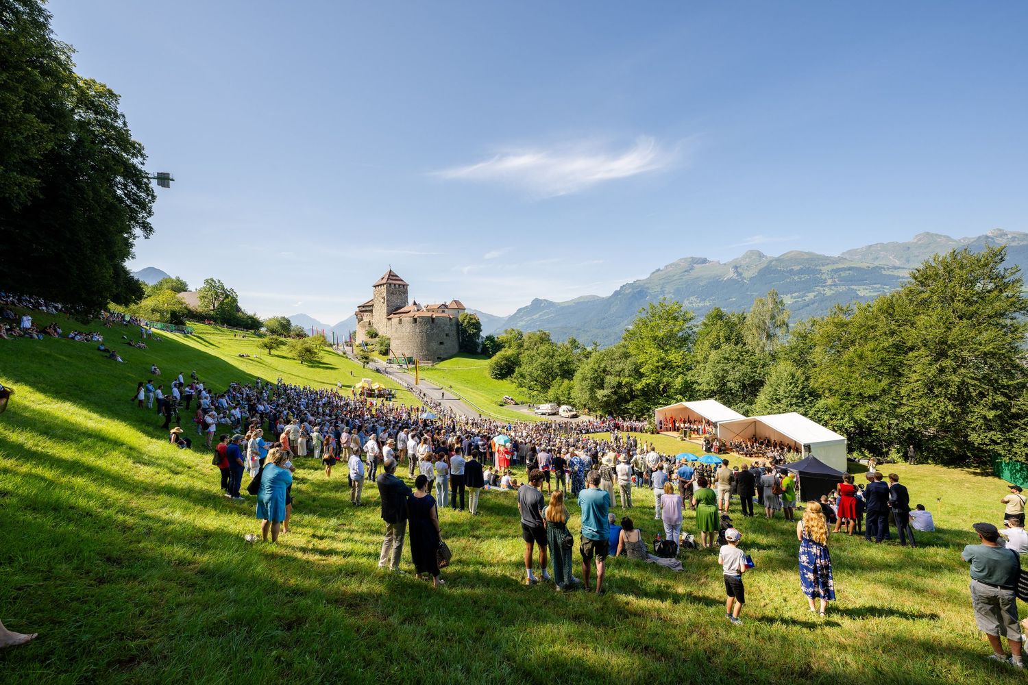 Crowd gathers at outdoor festival on green hillside with historic church and mountain backdrop in summer.