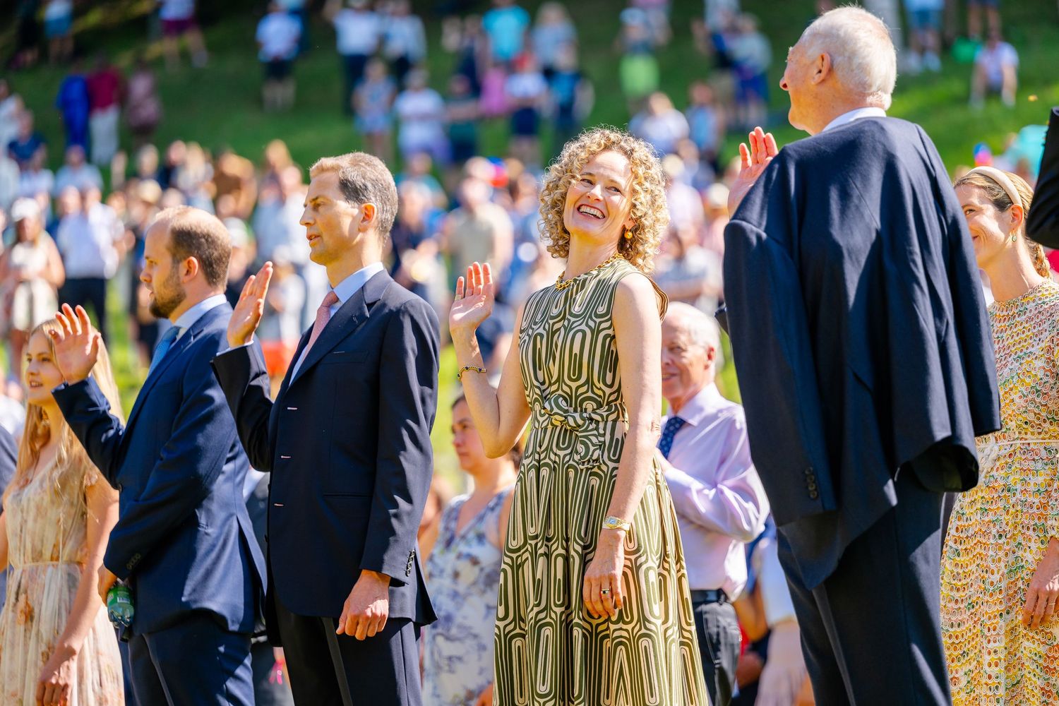 People attending an outdoor celebration event in formal attire on a sunny day with blurred crowd in background.