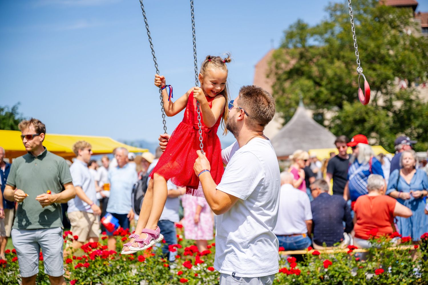 A child in a red dress enjoys swinging at an outdoor festival as an adult assists on a sunny summer day.