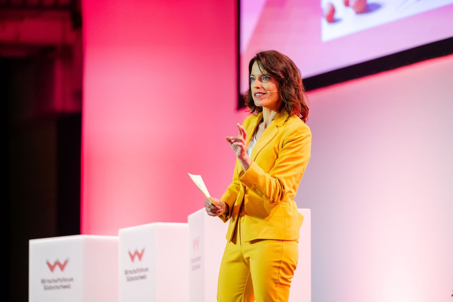 Professional speaker in bright yellow suit delivers engaging presentation on stage at conference event.