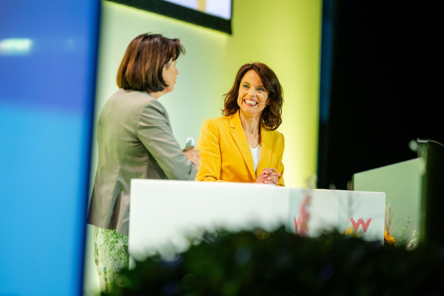 Conference speaker in yellow blazer engages with attendee at podium during networking session.