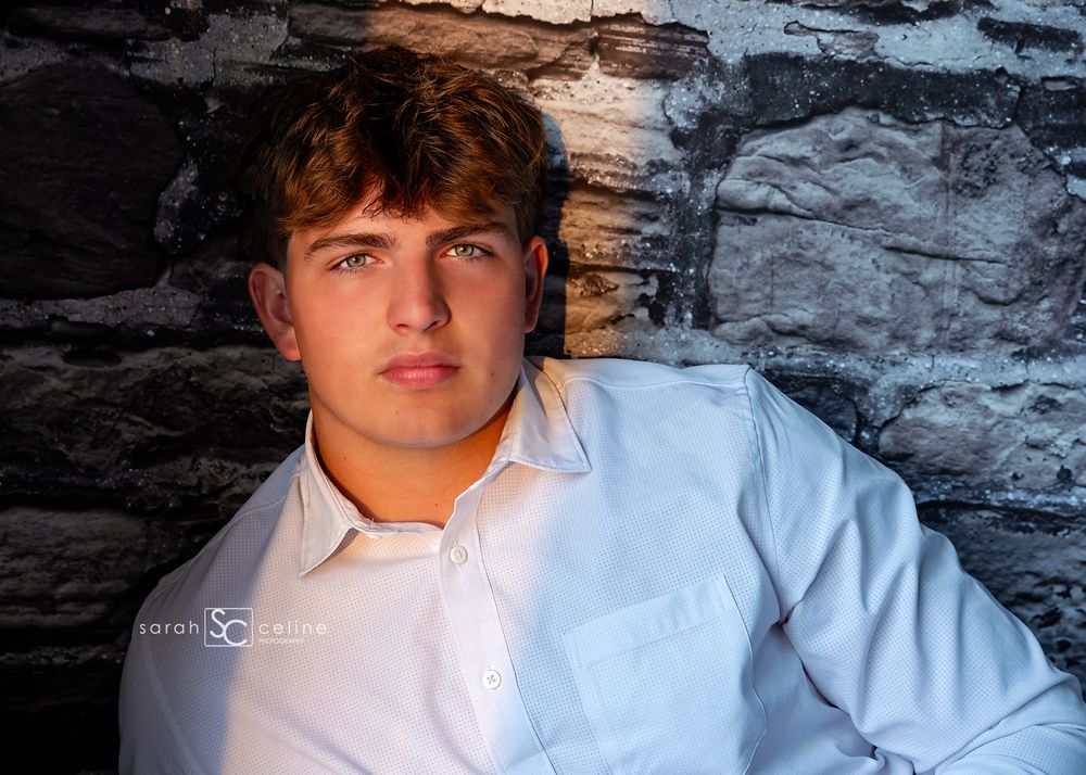 Young man in white shirt poses against stone wall in professional senior portrait photography.