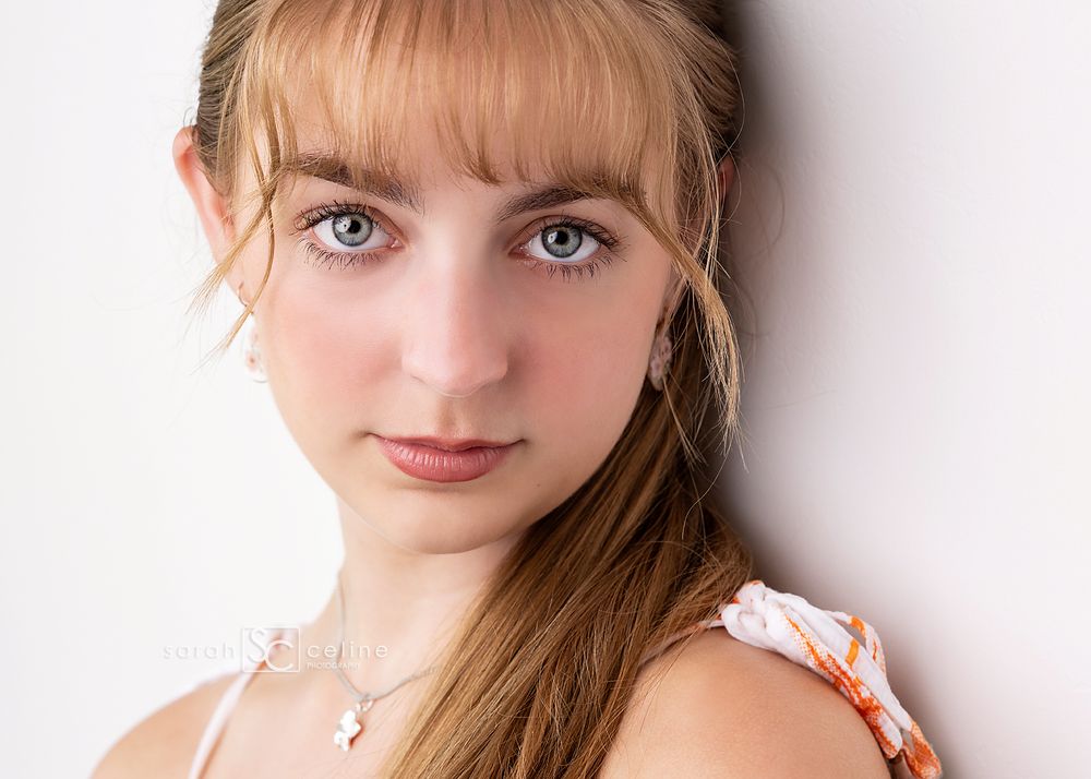 Young woman with blue eyes and long auburn hair wearing a floral top and necklace poses for a portrait.