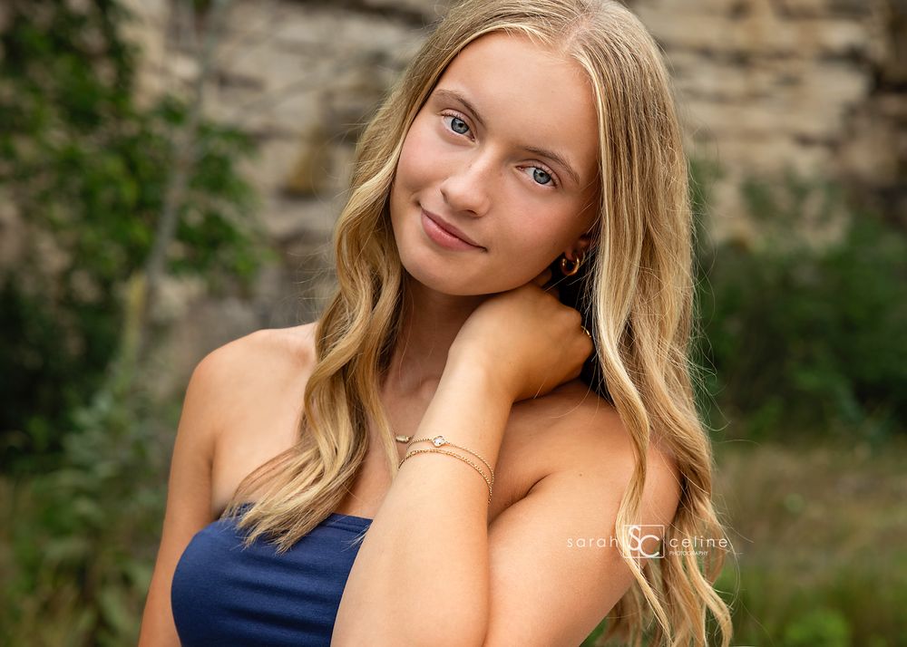 A young blonde woman in a navy top poses gracefully outdoors with a soft smile and bracelet.