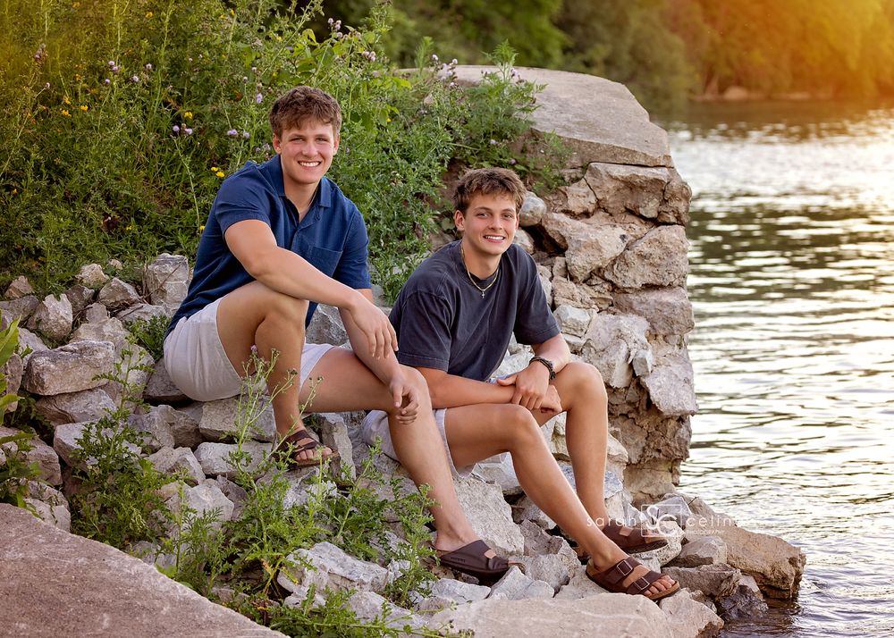 Two teenage boys smiling and sitting on rocky shoreline near a river, wearing casual summer clothes outdoors.