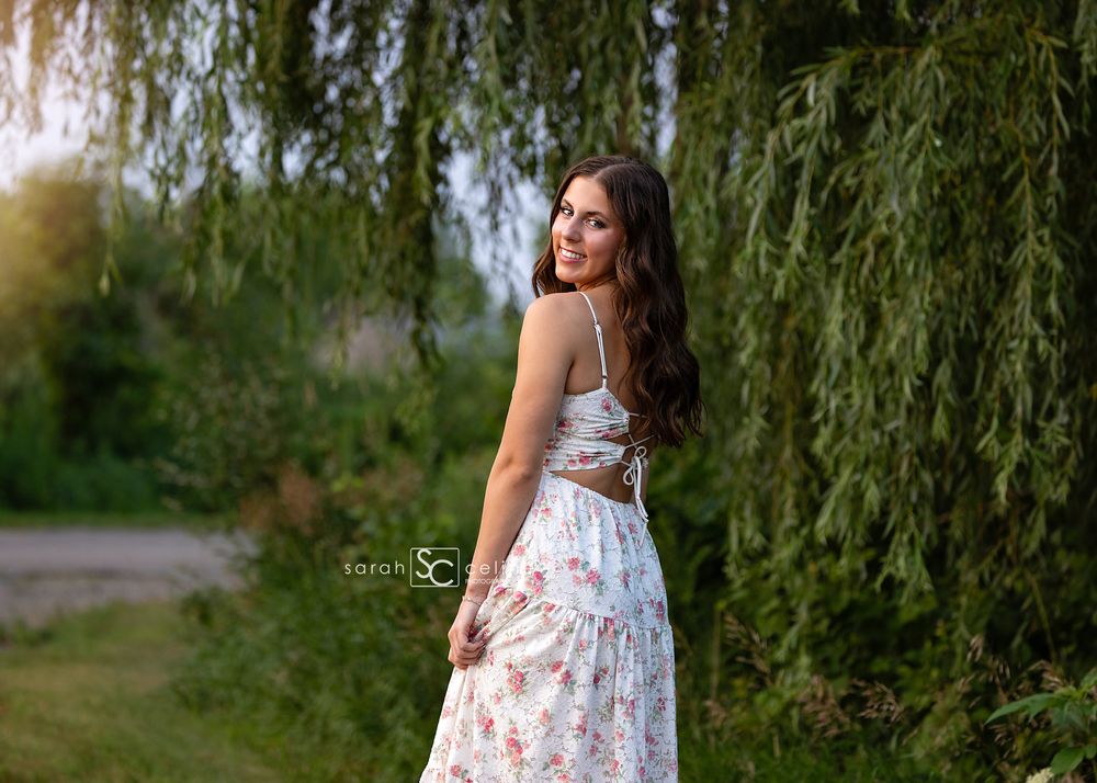 Young woman in floral maxi dress poses outdoors near weeping willow trees in golden hour sunlight.