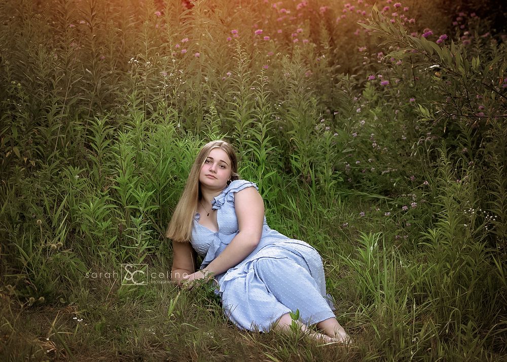 Young woman in blue dress sitting gracefully in a lush green wildflower meadow during golden hour.