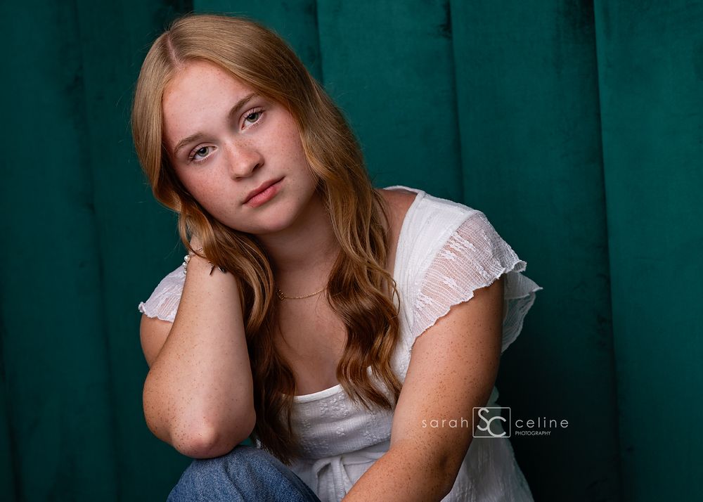A teenage girl with long auburn hair poses gracefully against a teal backdrop in a white ruffled top.