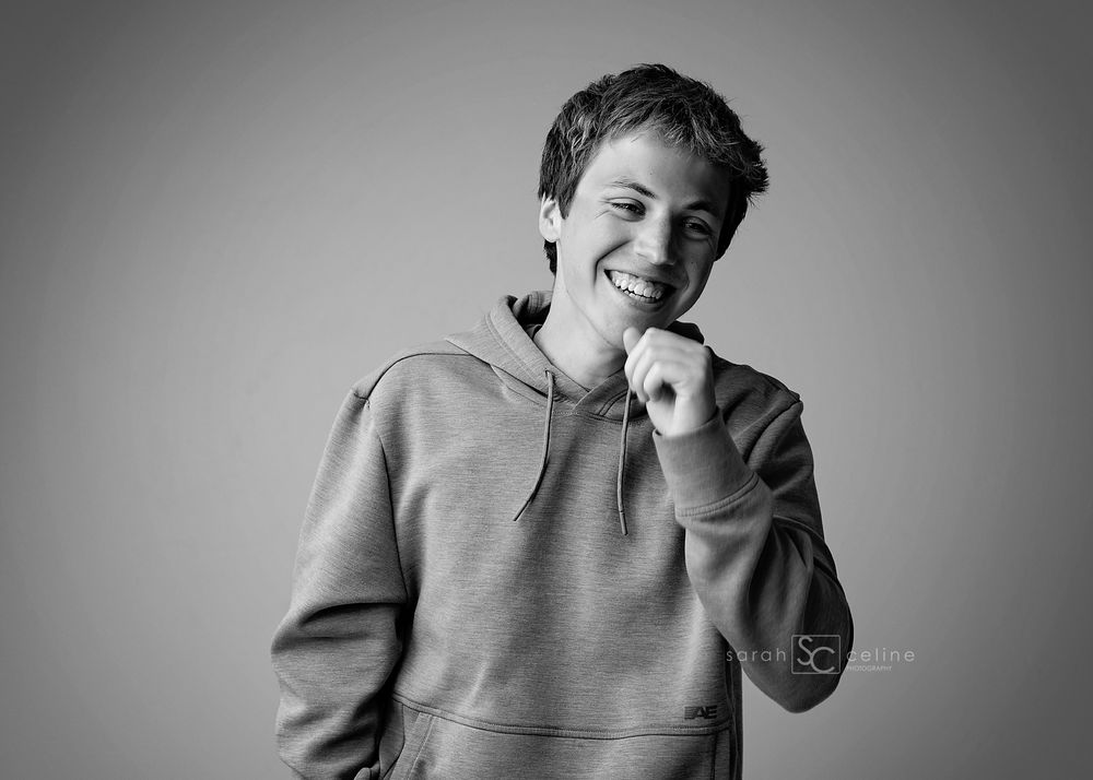 Black and white portrait of a smiling young man in a hoodie posing confidently against a gray background.