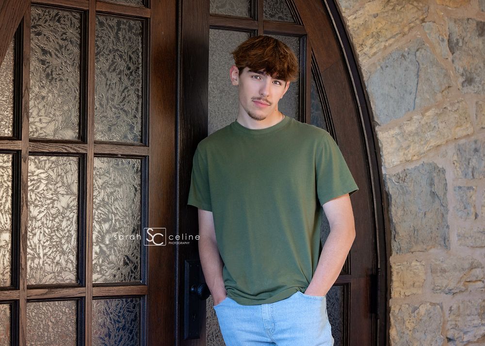 Young man in olive green t-shirt and light jeans posing confidently by ornate wooden door against stone wall.