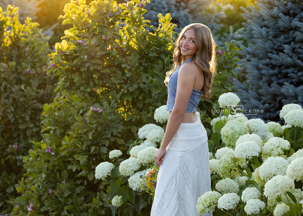 Smiling blonde woman in white skirt poses among white hydrangeas in a sunlit garden.