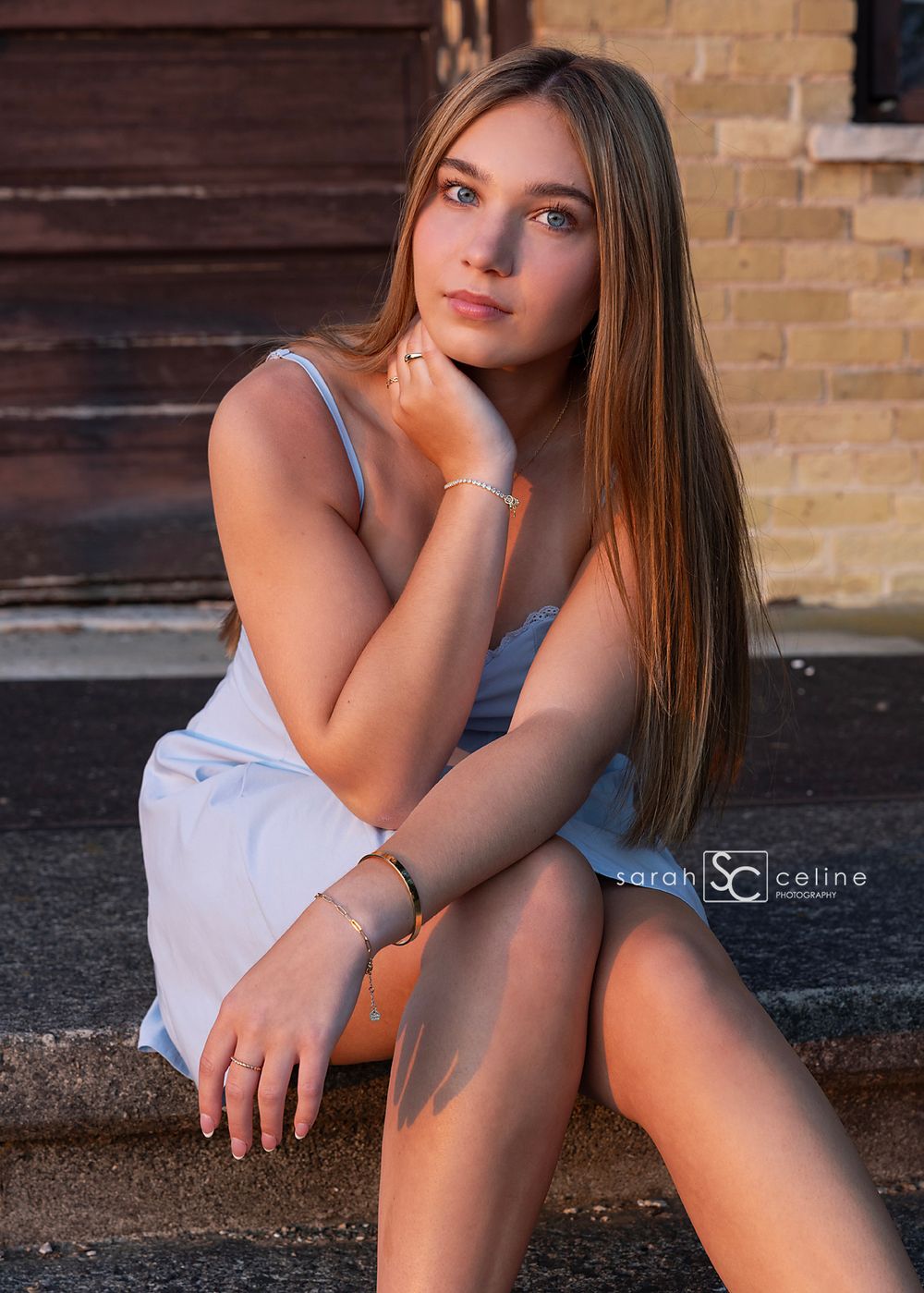 Young woman in a light blue dress sitting on steps, posing gracefully in golden hour sunlight.
