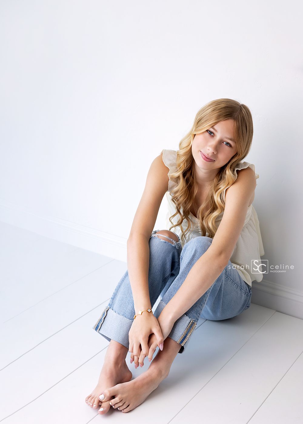 Young blonde woman in white tank top and ripped jeans sitting on white floor, smiling at camera.