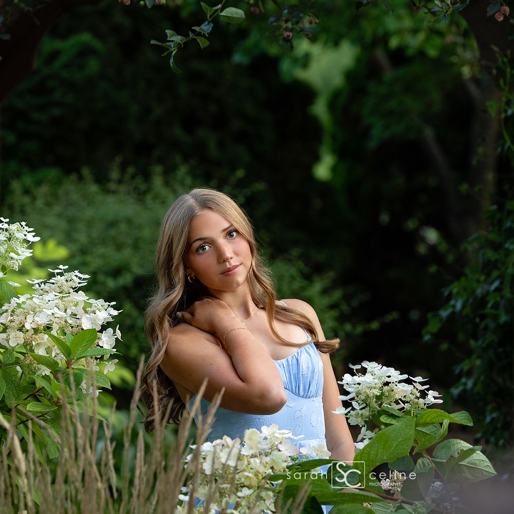 Young woman in light blue dress poses among white hydrangeas in a lush green garden setting.