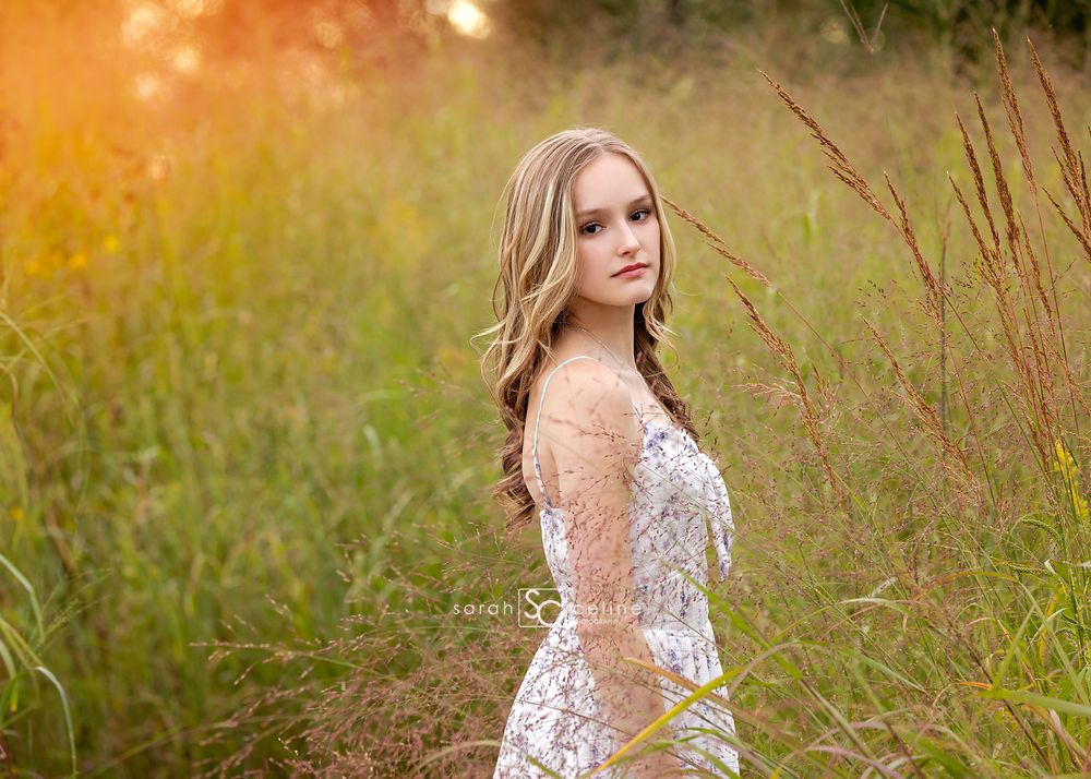 Young woman in white lace dress poses gracefully in a sunlit golden field with tall green grasses.