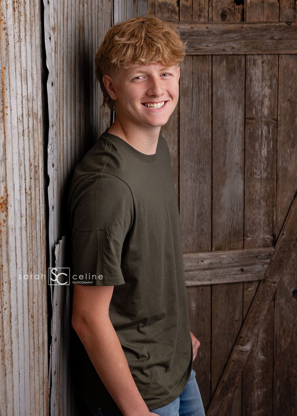 Smiling teenage boy with curly red hair wearing olive green t-shirt and jeans, leaning against rustic wooden barn door.