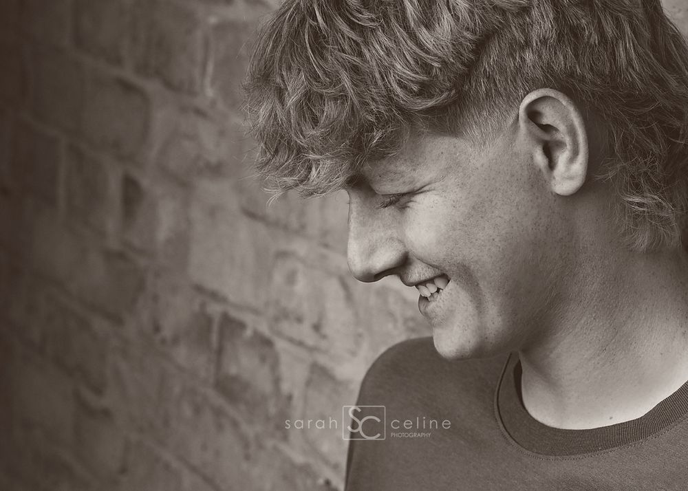 Smiling young man with curly hair laughing against a brick wall in a black and white portrait photo.