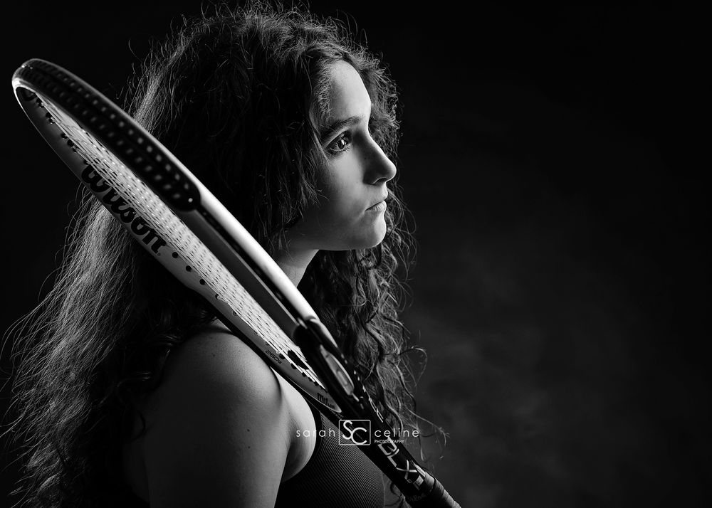 Black and white portrait of a young woman with curly hair holding tennis rackets over her shoulder.