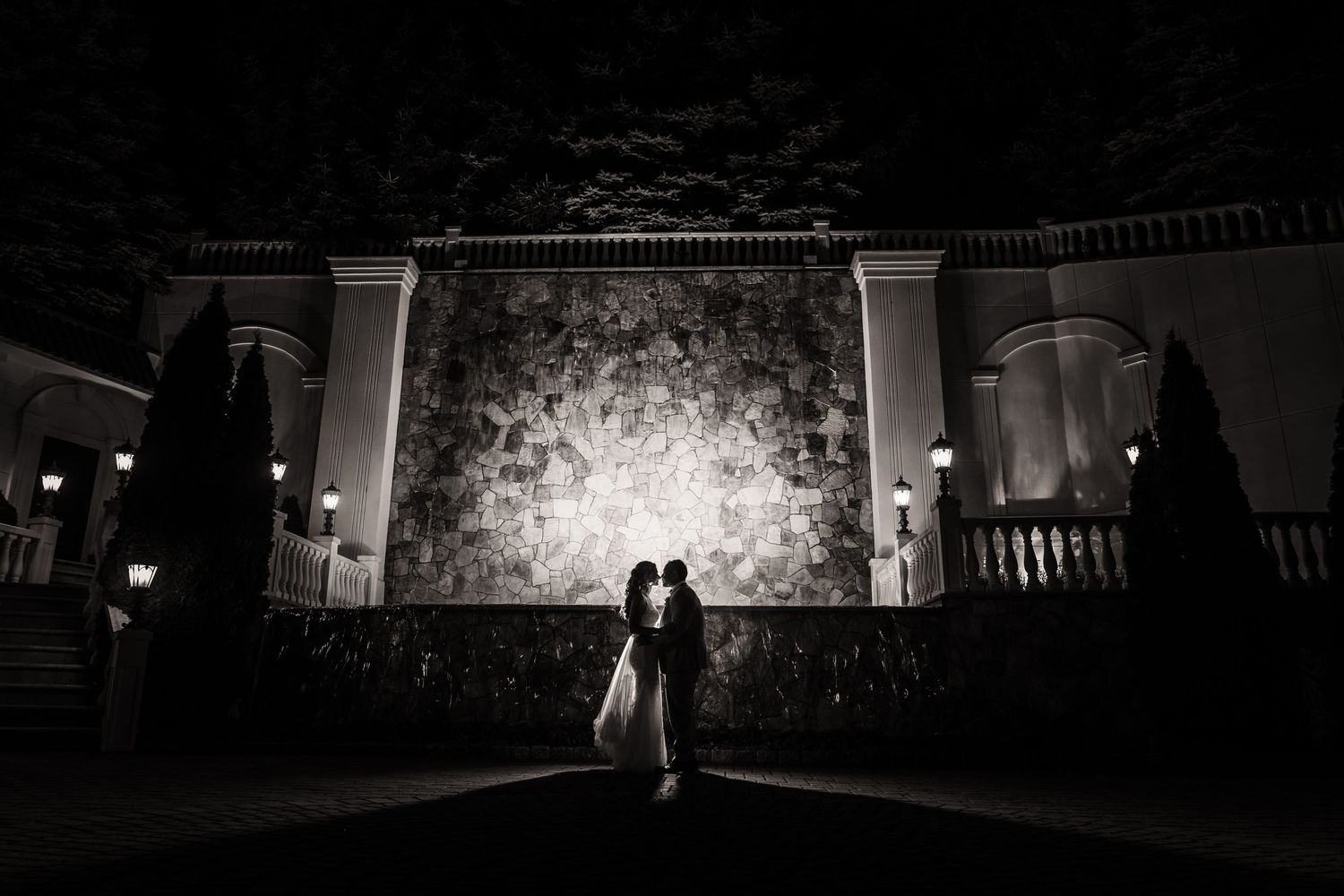 Two silhouettes stand against a brightly lit garden wall at night, creating a romantic black and white wedding scene.