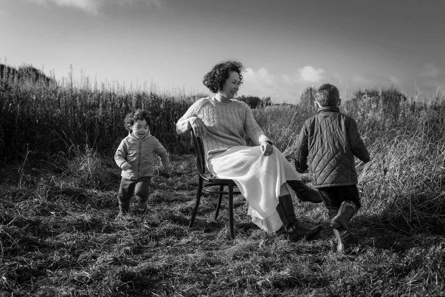 Black and white photograph of a mother seated outdoors while her young children move around her in tall grass