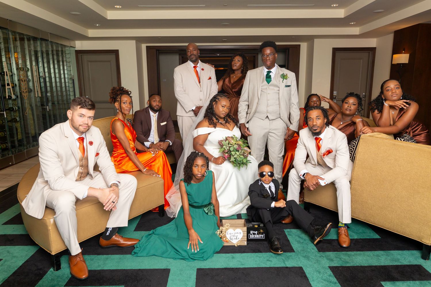 Wedding party in white tuxedos and colorful dresses pose together on emerald green carpeted floor in elegant room.