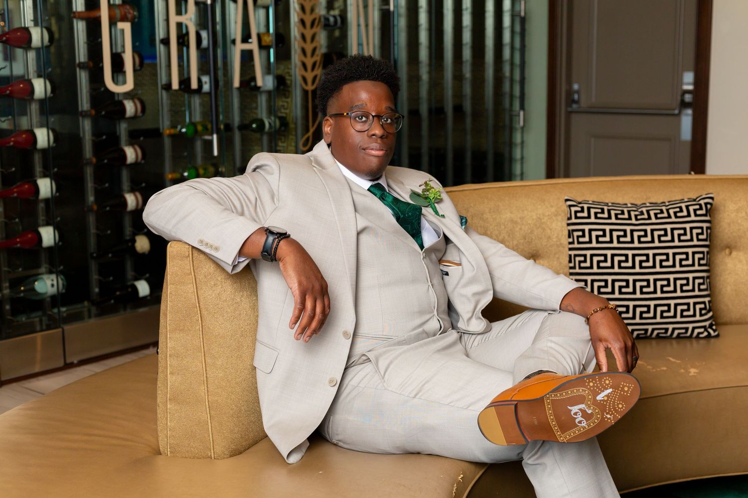 A person relaxes on a tan couch wearing a light-colored suit and brown dress shoes in a well-lit modern space.