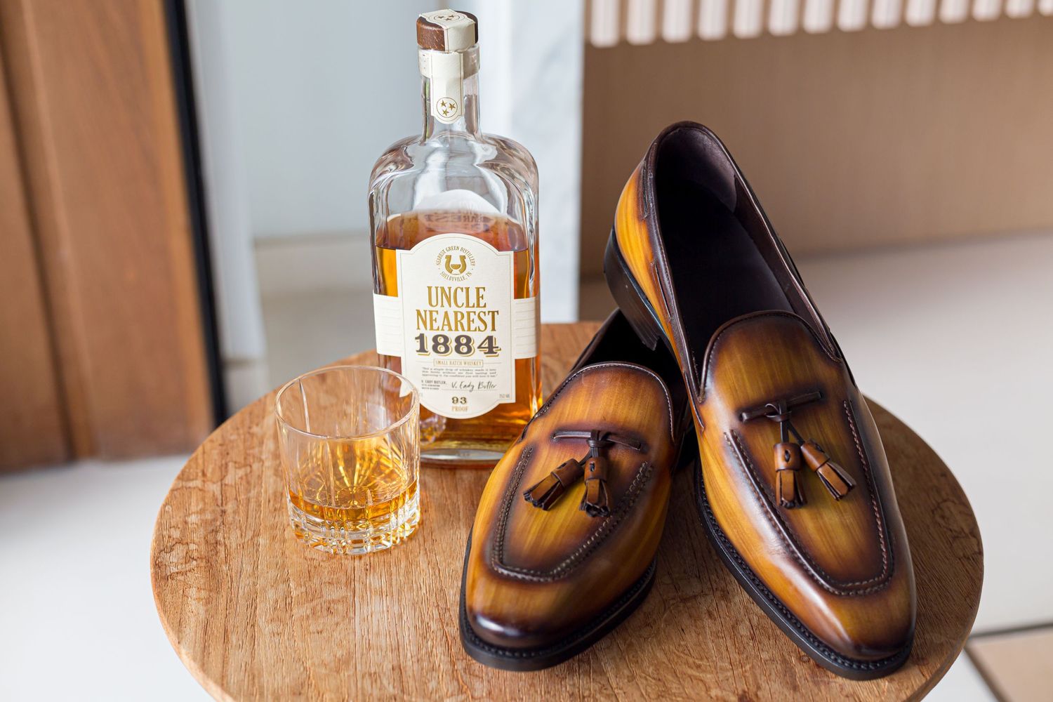 Elegant brown leather tassel loafers displayed next to whiskey bottle and glass on wooden surface.