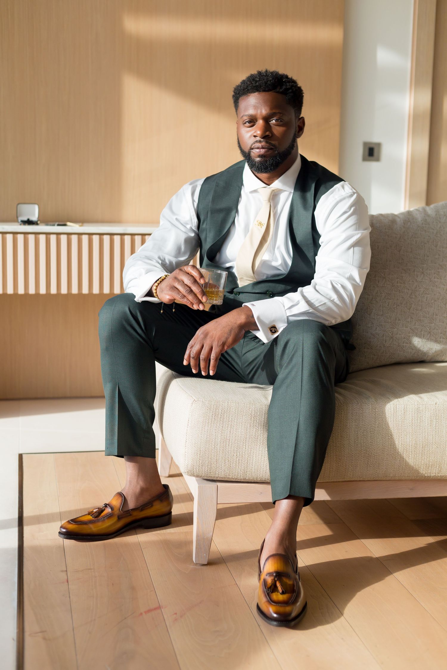 A well-dressed person in a green vest and white shirt sits on a beige couch wearing tan leather loafers.