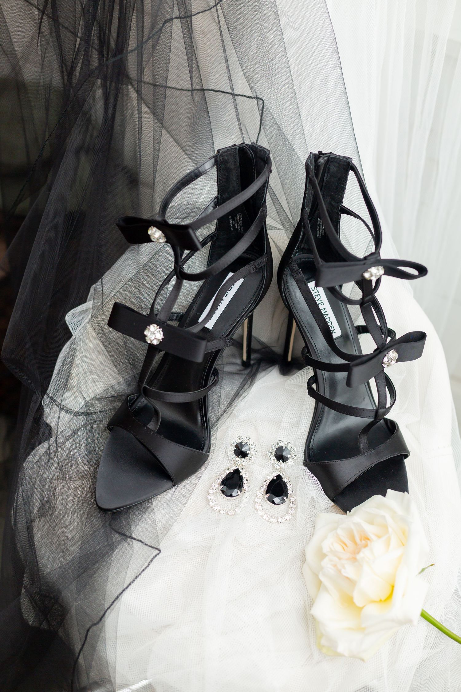 Black strappy wedding shoes with crystal earrings and white rose on tulle fabric.