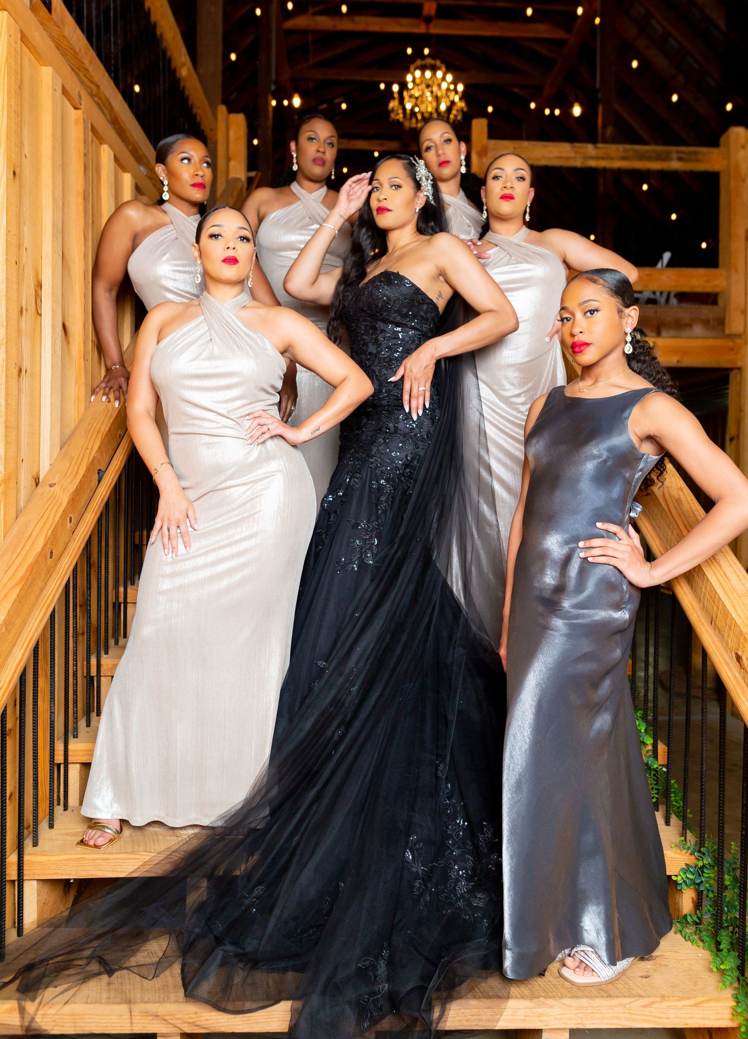 A glamorous group in formal attire poses on a wooden staircase, with one in a black gown surrounded by silver dresses.