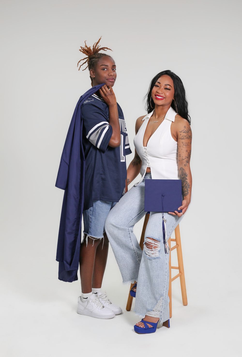 Two stylish women pose together, one in a navy graduation gown and jersey, the other in white halter top and ripped jeans.