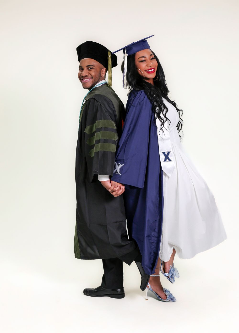 Two graduates in caps and gowns, one in black doctoral robes and one in blue, pose back-to-back holding hands.