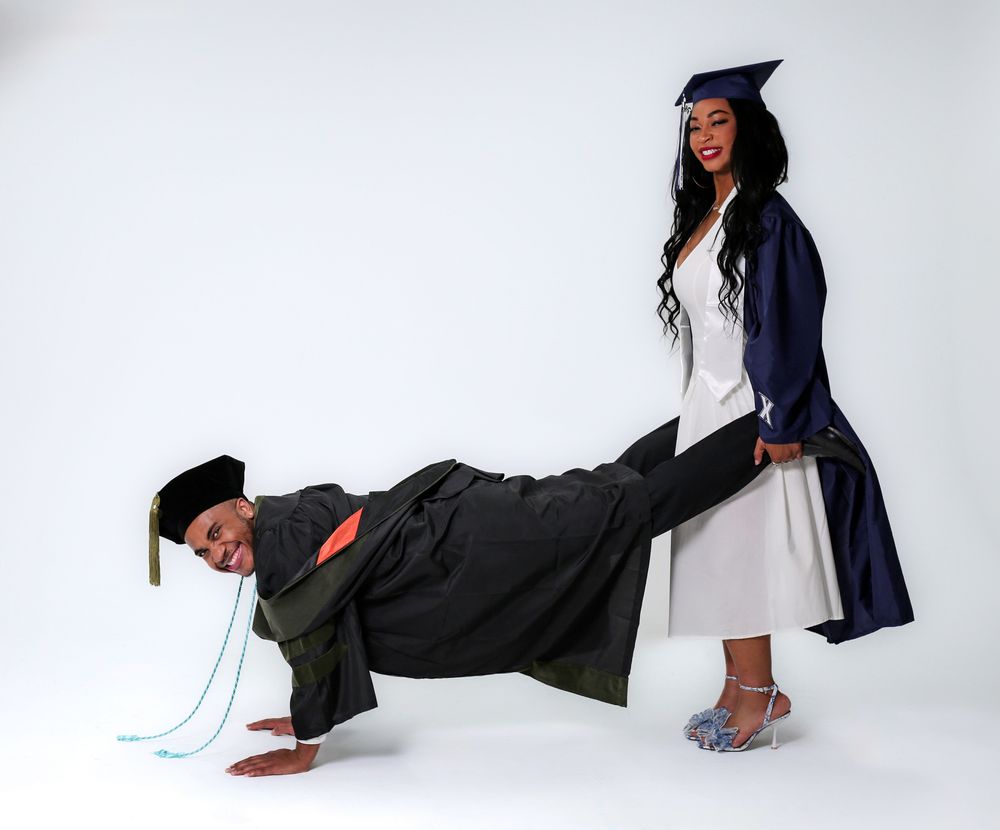 Two graduates in caps and gowns pose playfully, one doing a plank while the other stands on their back.