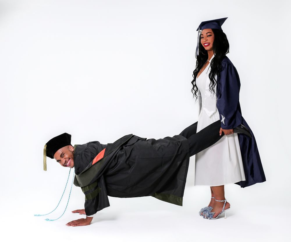 Two graduates in caps and gowns pose playfully, one doing a plank while the other stands smiling in white dress.