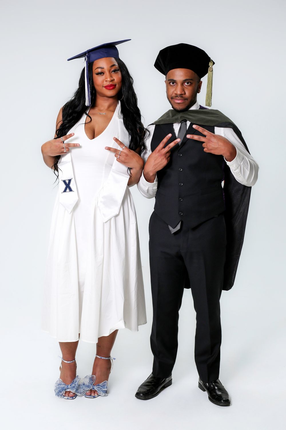 Two graduates in caps and gowns posing together, woman in white dress and man in black suit, celebrating their achievement.